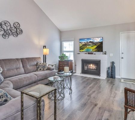 Cozy living room with gray sectional sofa, glass and metal tables, fireplace, TV, wooden floor, and neutral wall decor.