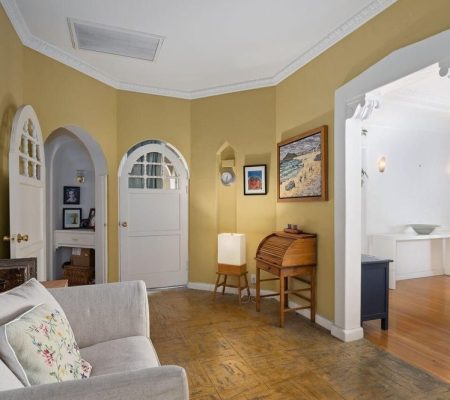 Cozy living room with beige walls, arched doorways, a light gray sofa, wooden roll-top desk, and wall art near a bright adjacent room.