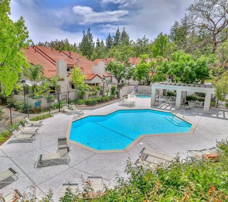 Outdoor swimming pool surrounded by lounge chairs, trees, and red-roofed residential buildings under a cloudy sky.