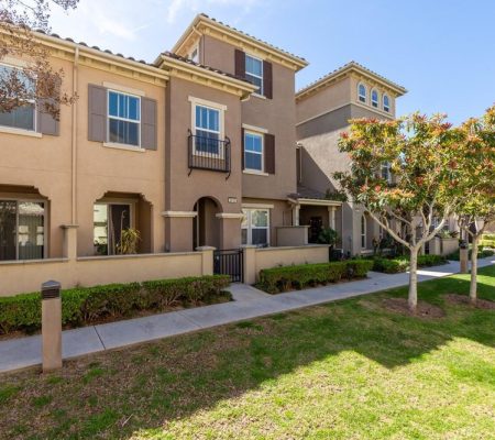 Exterior view of a beige townhouse with a small front yard, sidewalk, green grass, and several trees under a clear blue sky.