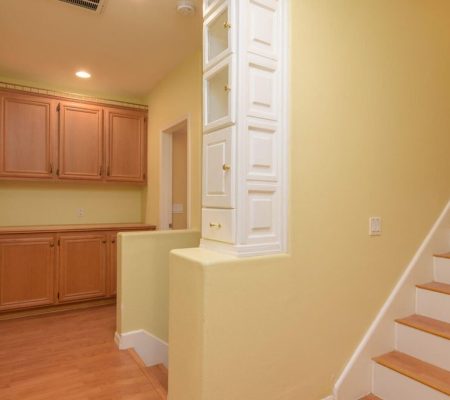 Interior view of a hallway with wooden cabinets, a round wall mirror, white stairs with wood trim, and pale yellow walls.